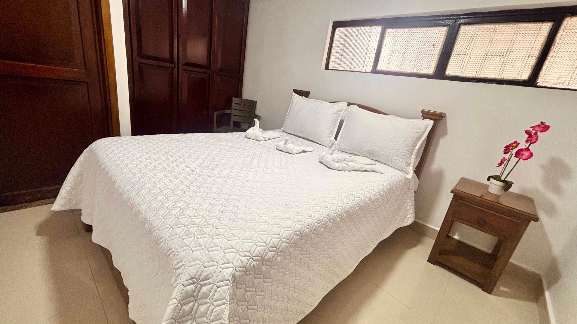 Cama bien hecha en un dormitorio minimalista con muebles de madera y un pequeño jarrón con flores rosas.