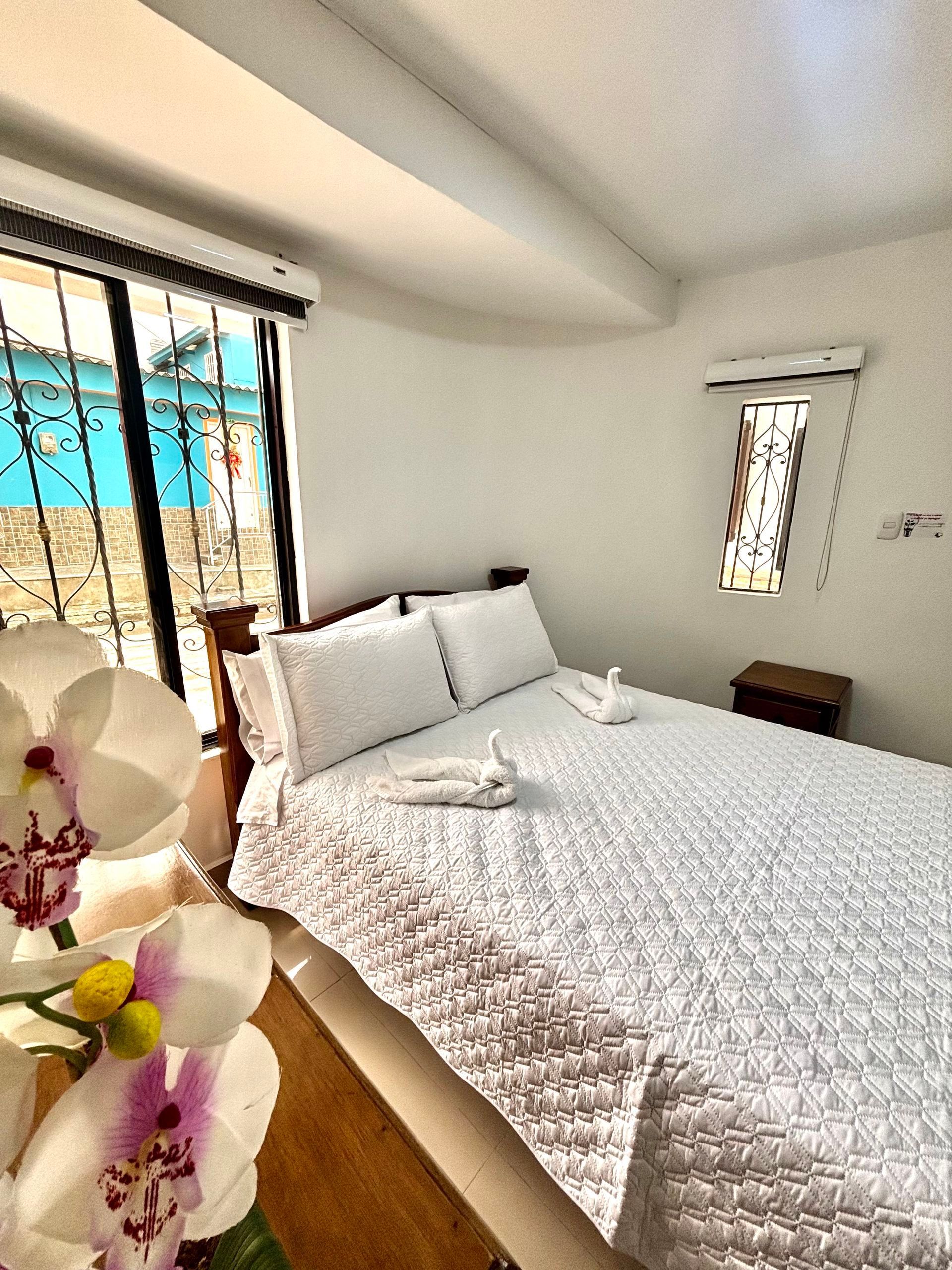 Habitación luminosa con ropa de cama blanca, puertas corredizas de vidrio y un jarrón de orquídeas en primer plano.