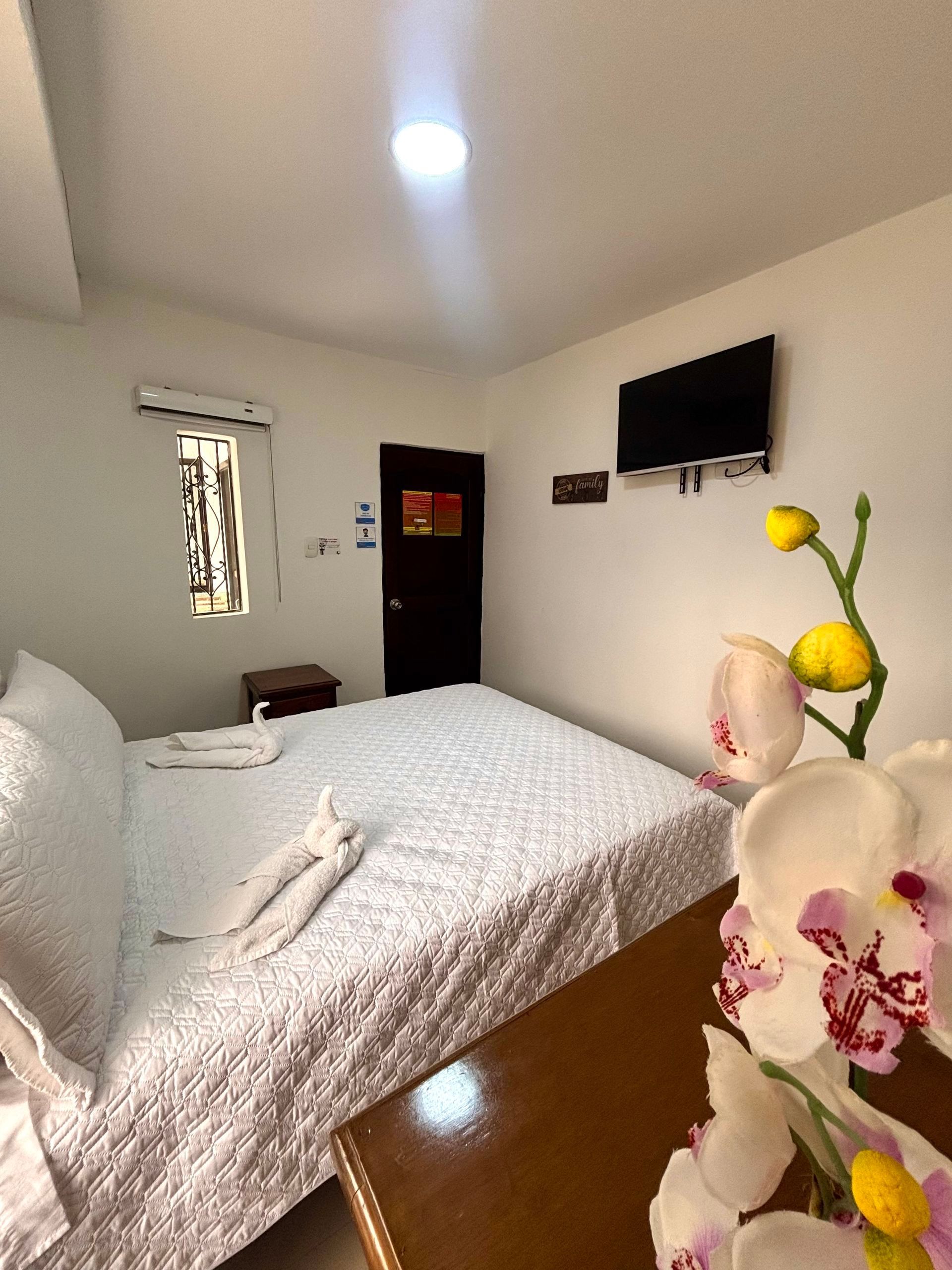 Habitación pequeña con una cama, un televisor montado en la pared y un arreglo floral en primer plano.