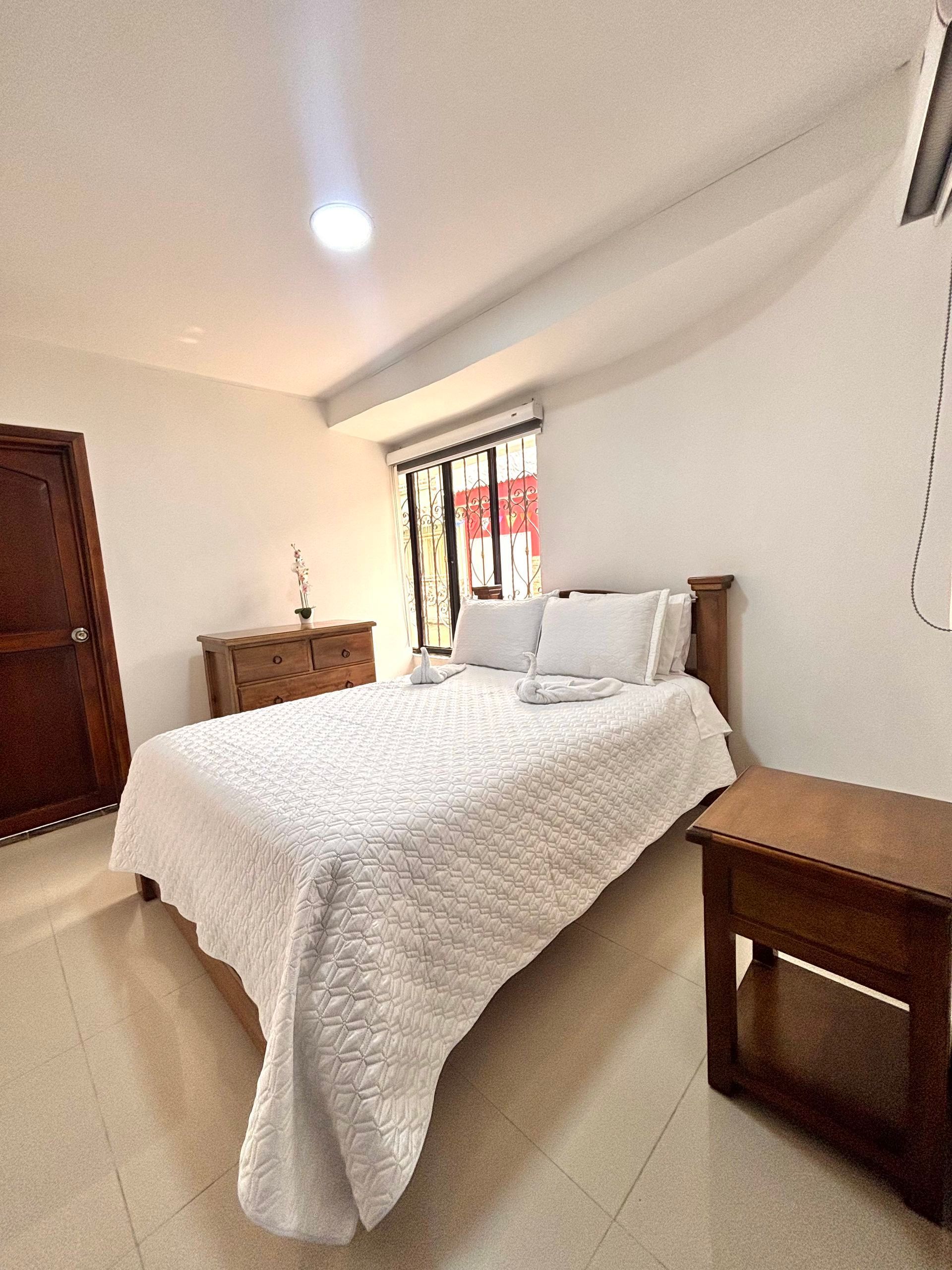 Habitación luminosa con cama blanca, muebles de madera y luz natural que entra por la ventana.