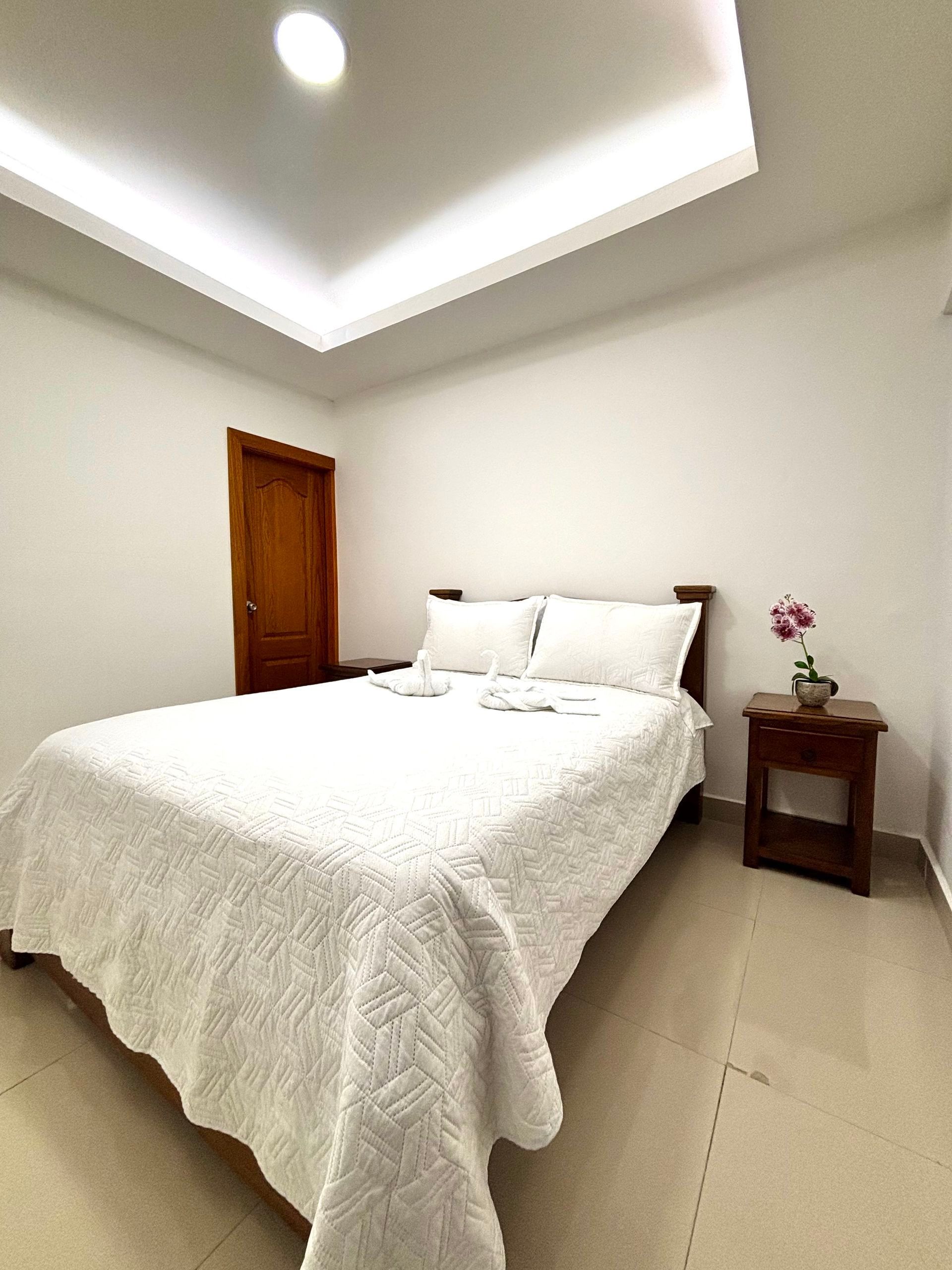Dormitorio luminoso y minimalista con cama blanca, puerta de madera y mesita de noche con flores.