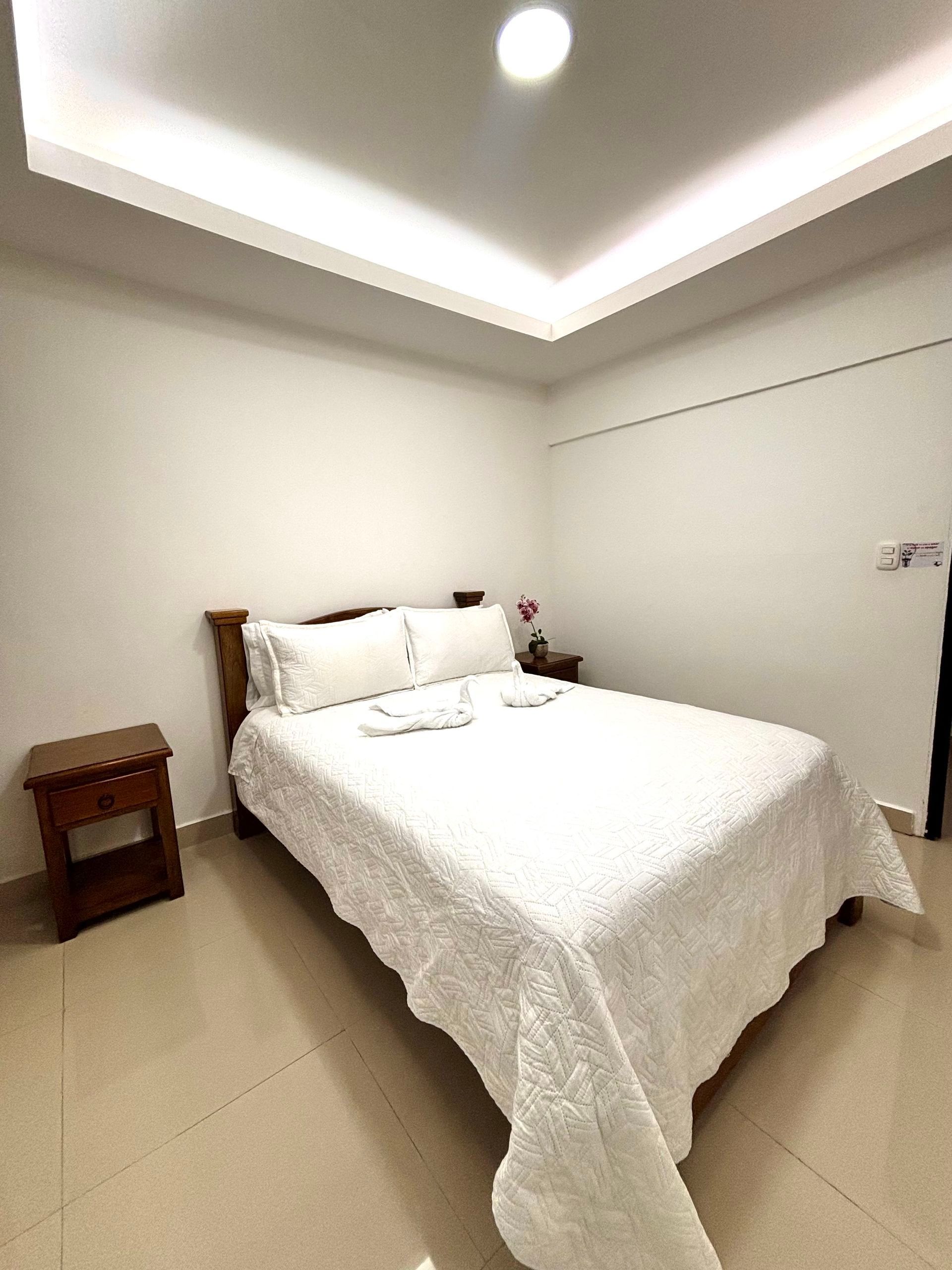 Dormitorio minimalista con una cama blanca, una pequeña mesita de noche de madera y luces empotradas en el techo.