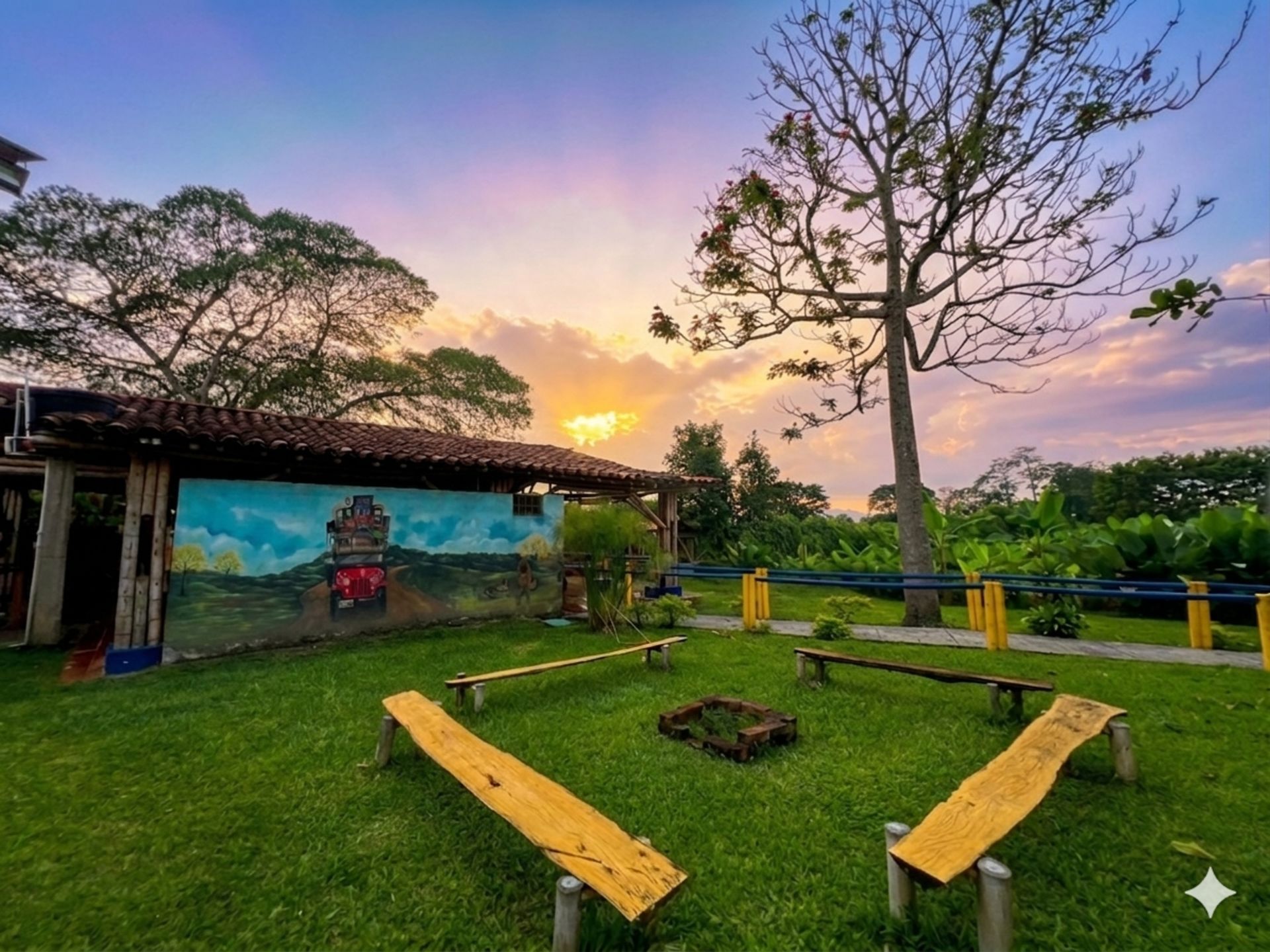 Puesta de sol sobre un patio cubierto de césped con bancos amarillos, un mural en la pared, árboles y cielo azul.