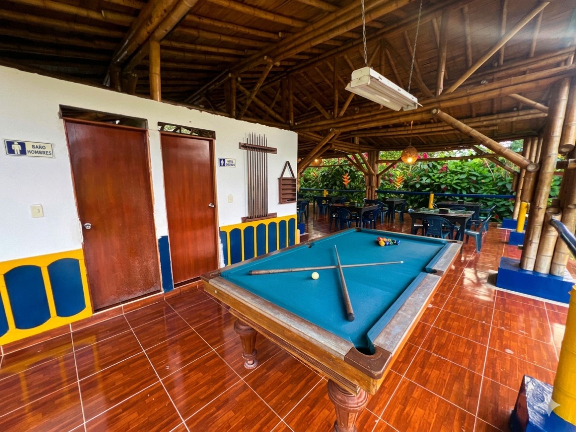Sala de billar cubierta con una mesa de billar azul, suelo de madera y asientos al aire libre al fondo.