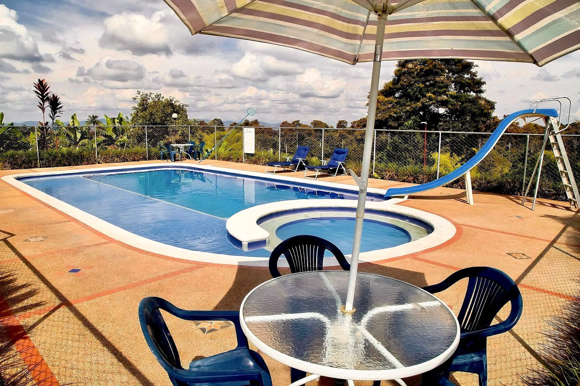 Piscina en el patio trasero con tobogán azul, mesa y sillas de patio bajo una sombrilla a rayas.