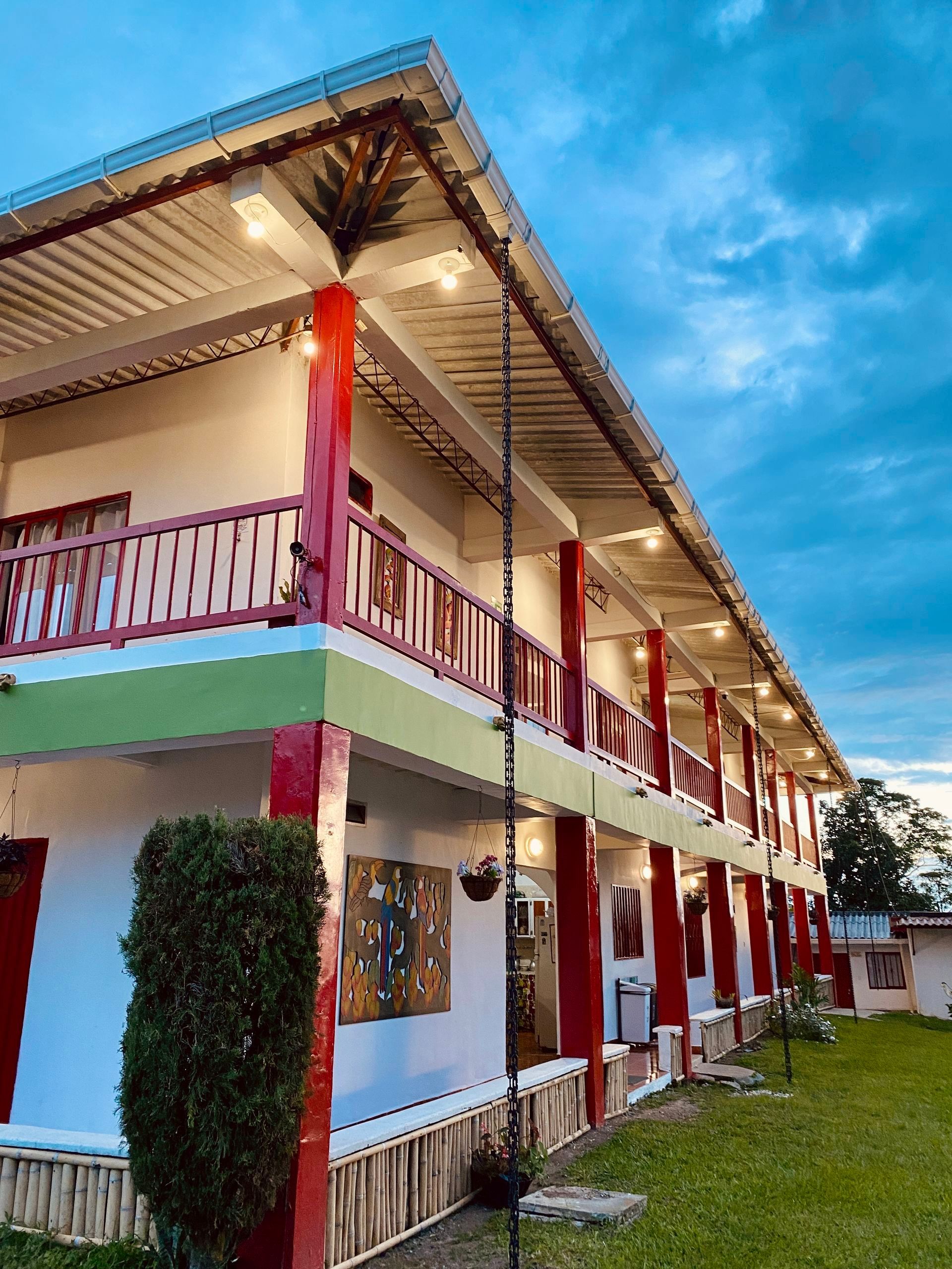 Edificio de dos plantas, blanco y rojo, con balcones junto a un césped bajo un cielo azul.