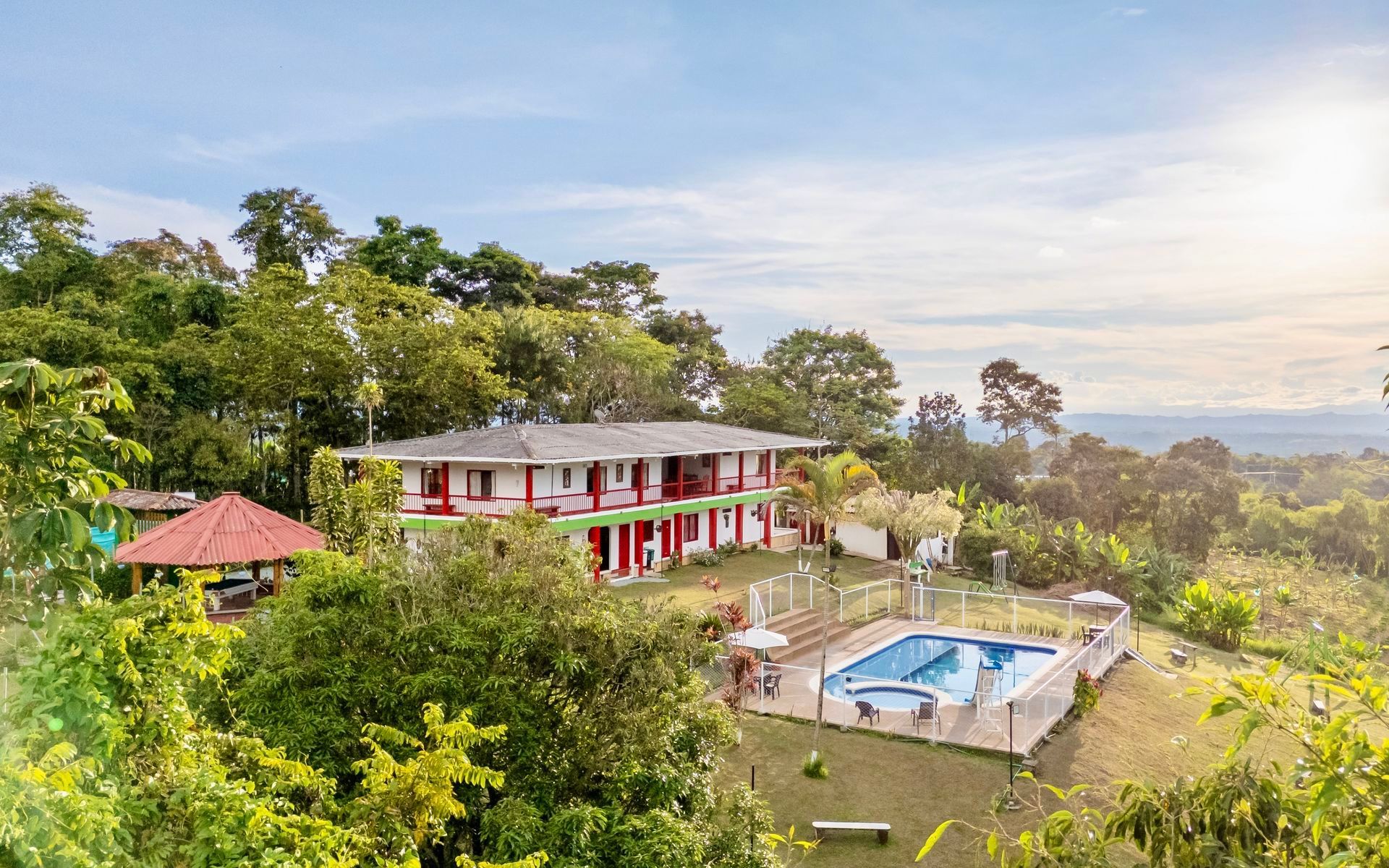 Villa en la cima de una colina con detalles en rojo, piscina y exuberantes árboles tropicales al atardecer.