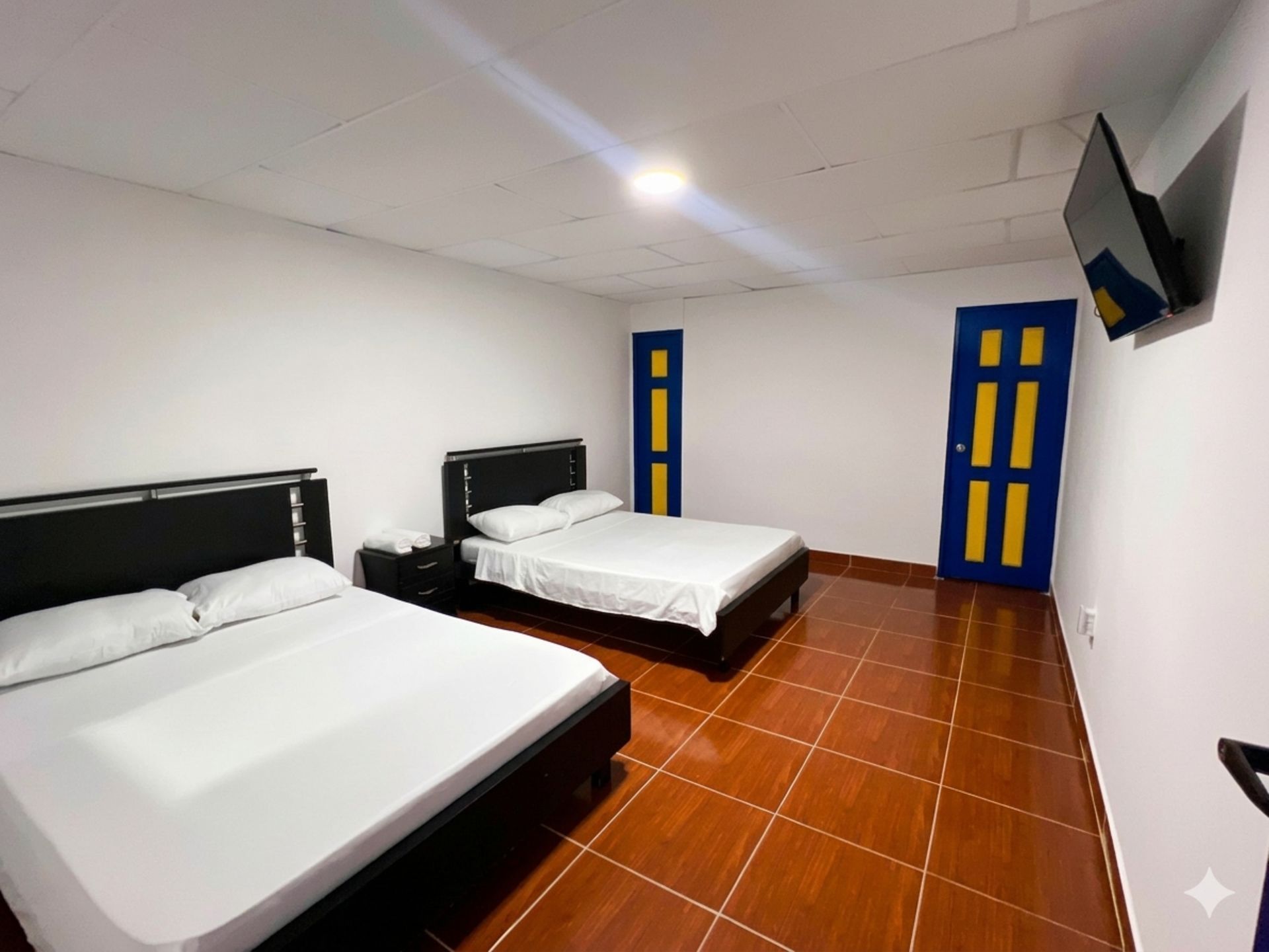 Habitación de hotel sencilla con dos camas, suelo de baldosas naranjas, puertas azules y un televisor montado en la pared.