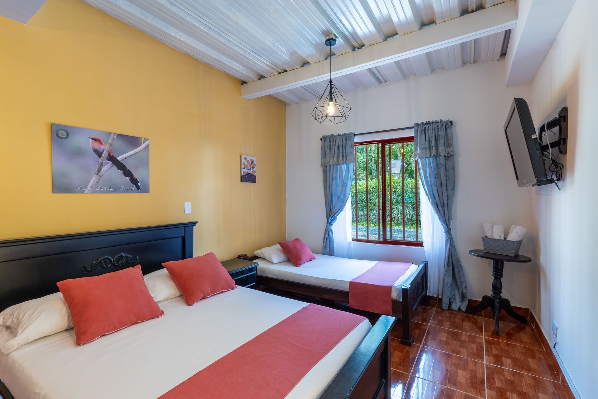 Habitación luminosa con dos camas, paredes amarillas, una ventana con cortinas azules y un televisor montado en la pared.