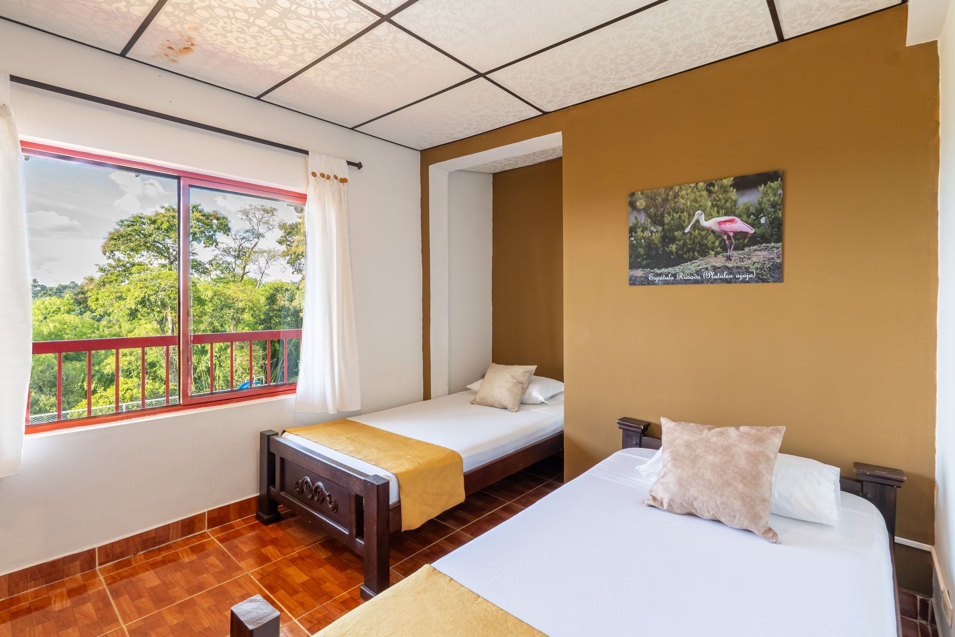 Habitación pequeña con dos camas individuales, vista a la ventana, cálido suelo de madera y pared decorativa.