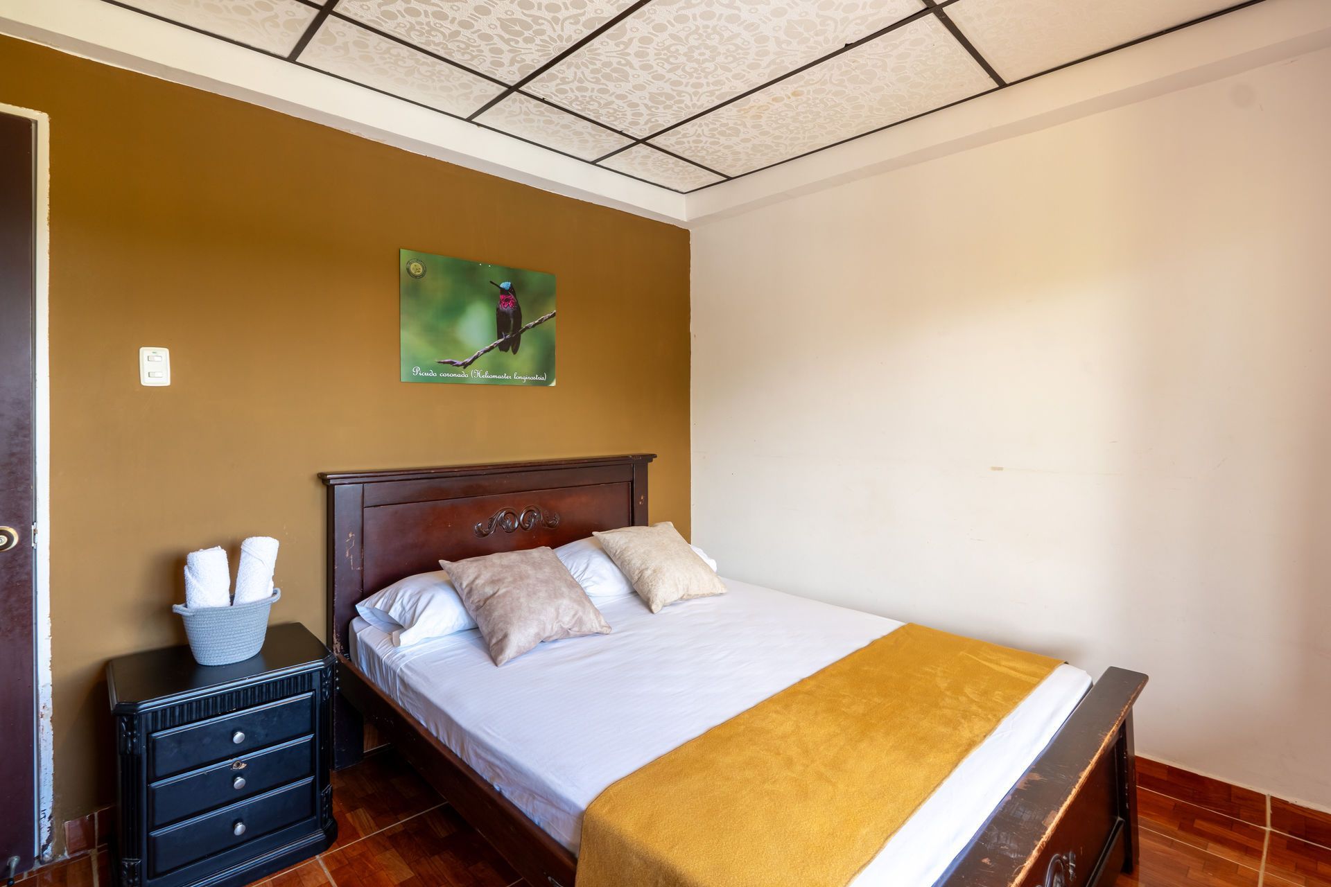 Dormitorio con cama de madera, pared decorativa color mostaza, pared beige y cómoda oscura junto a la cama.
