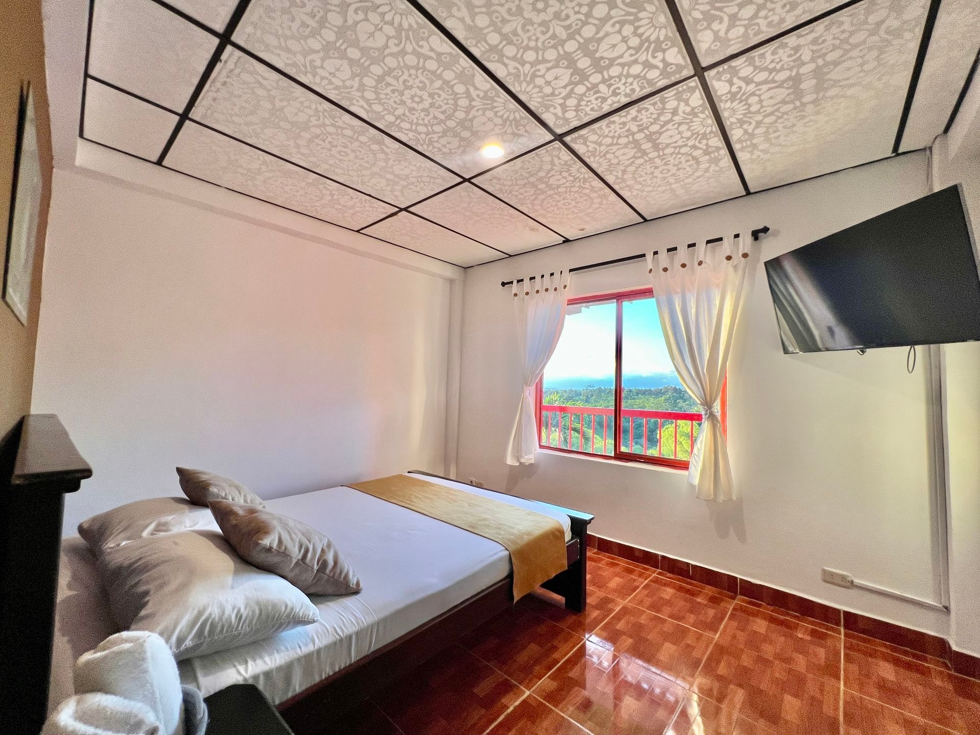 Habitación luminosa con cama, ventana, suelo de madera y televisor montado en la pared.