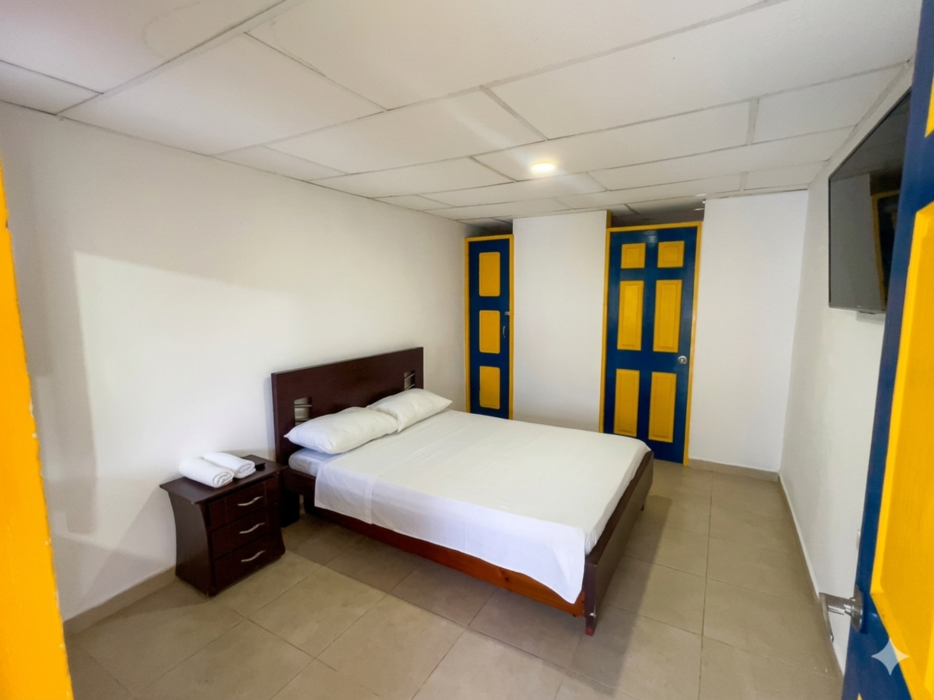 Habitación pequeña con cama, mesita de noche, paredes blancas y puertas azules y amarillas.