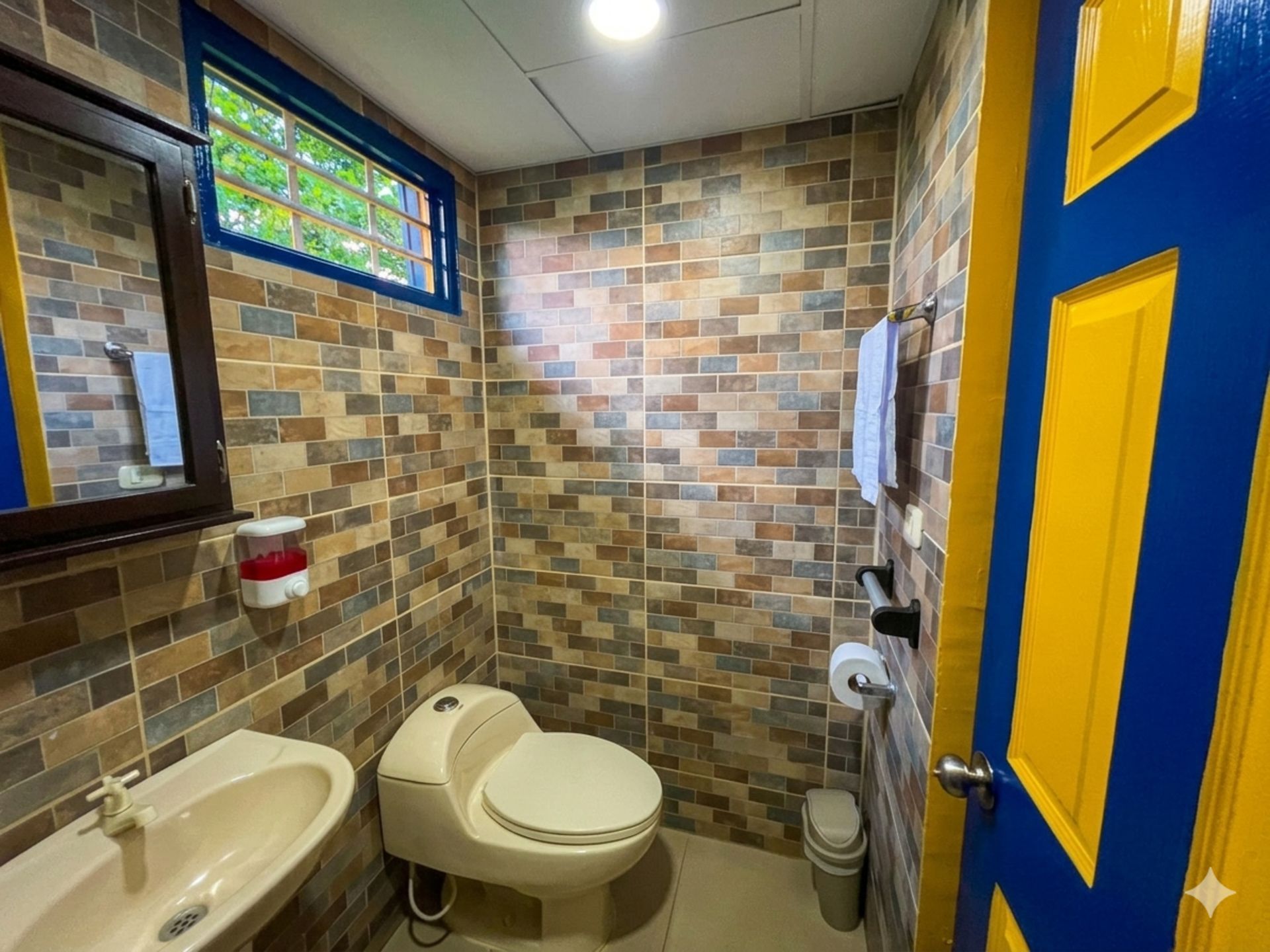 Pequeño baño alicatado con lavabo, inodoro y puerta azul y amarilla bajo una vidriera.