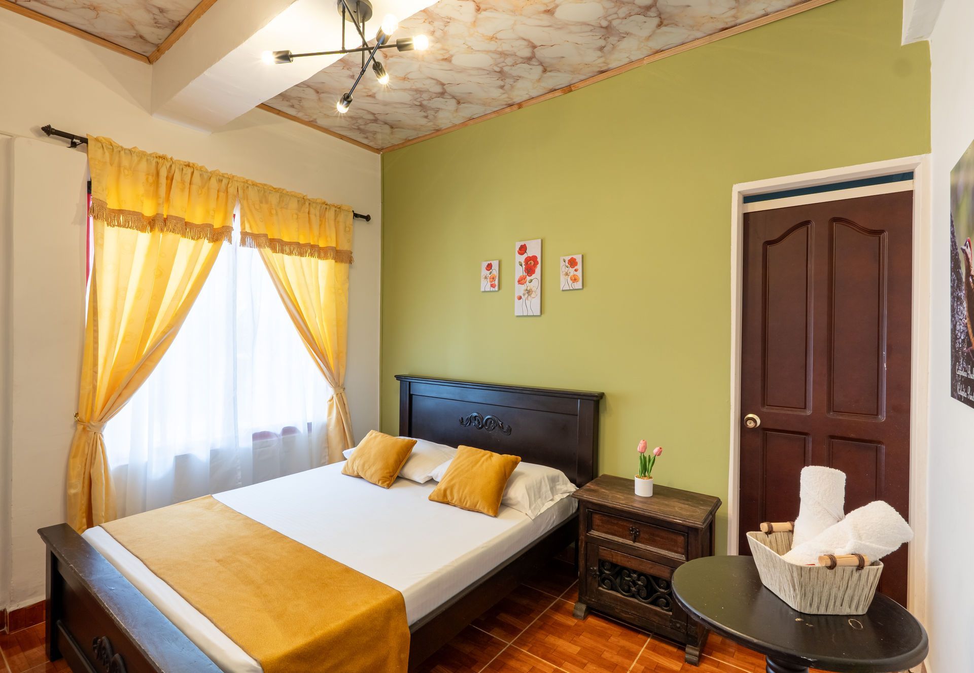 Habitación pequeña con una cama, cortinas amarillas, paredes verdes y una puerta de madera oscura.