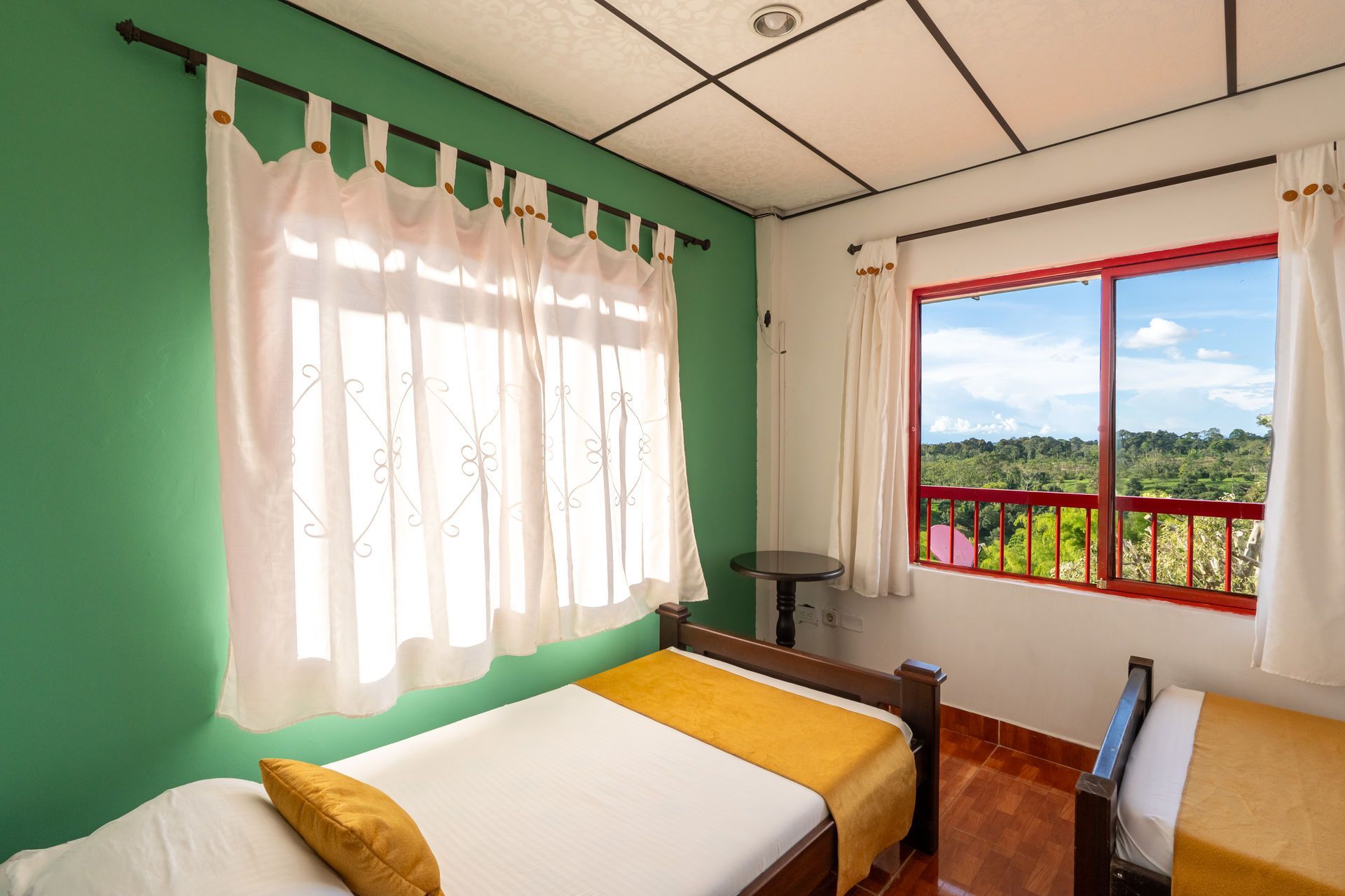 Habitación luminosa con paredes verdes, dos camas individuales y una ventana con vistas a las montañas y al cielo.