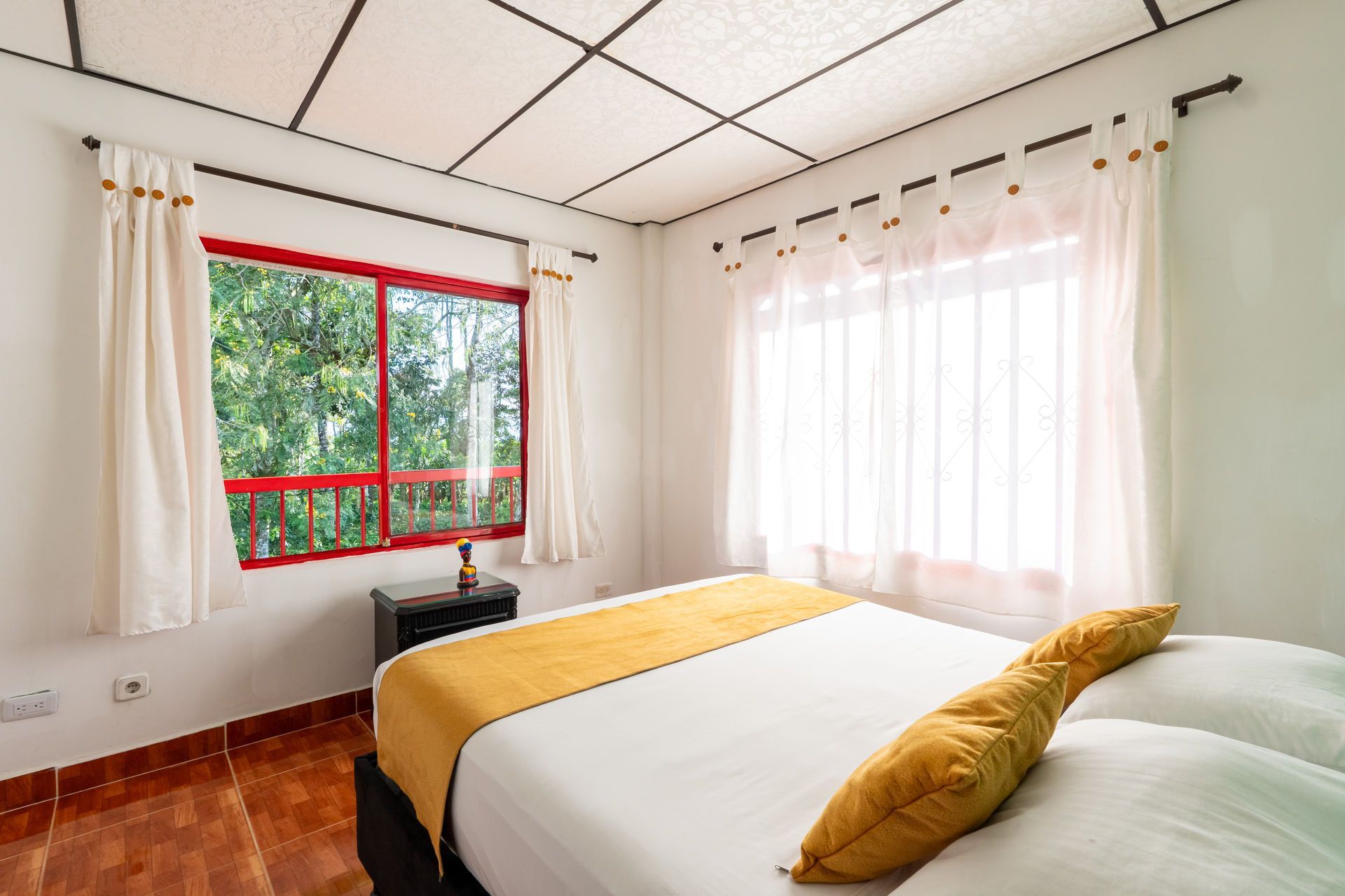 Habitación luminosa con cama, manta amarilla, grandes ventanales y cortinas blancas.