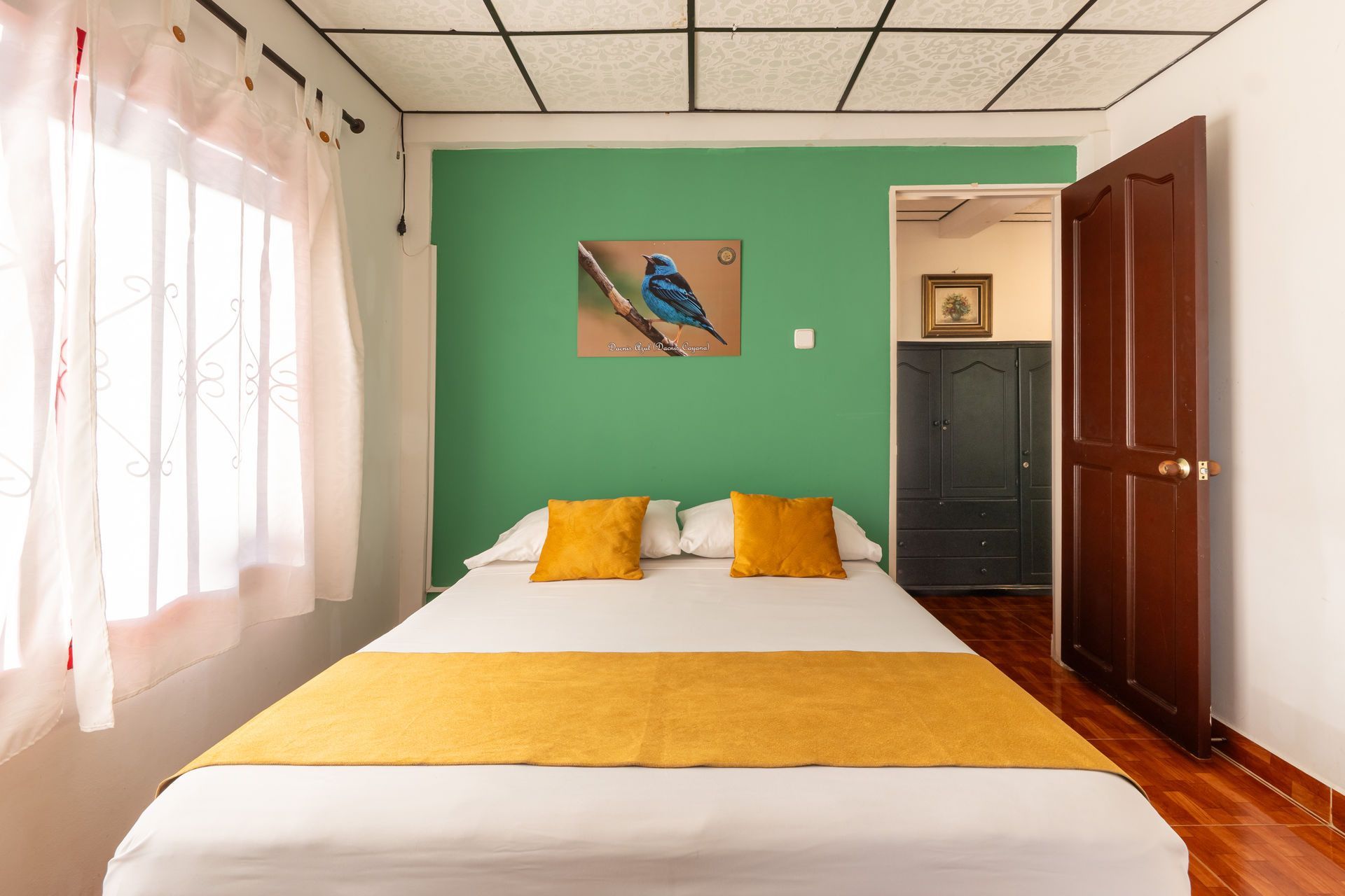 Habitación ordenada con cama, ropa de cama amarilla, pared decorativa verde, cortinas y puertas de madera abiertas.
