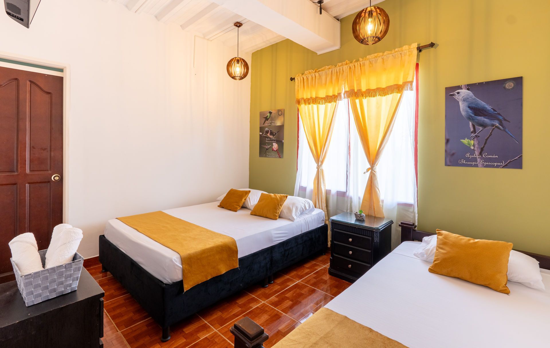 Habitación de hotel luminosa con dos camas individuales, cortinas amarillas, suelo de madera y cuadros en las paredes.
