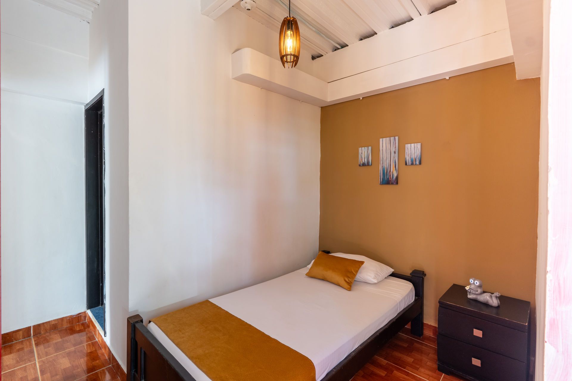 Habitación pequeña con cama, pared decorativa de color naranja, mesita de noche y lámparas colgantes.