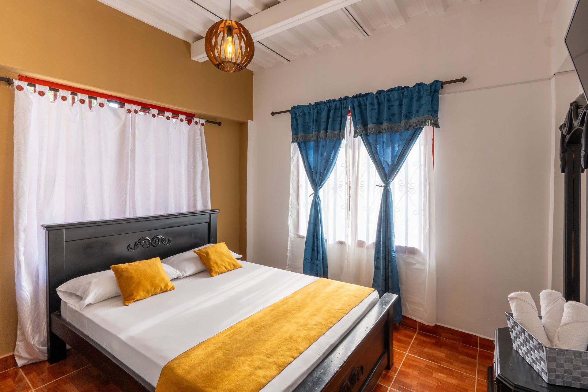 Habitación acogedora con cama negra, ropa de cama blanca, camino de mesa amarillo y cortinas azules junto a la ventana.