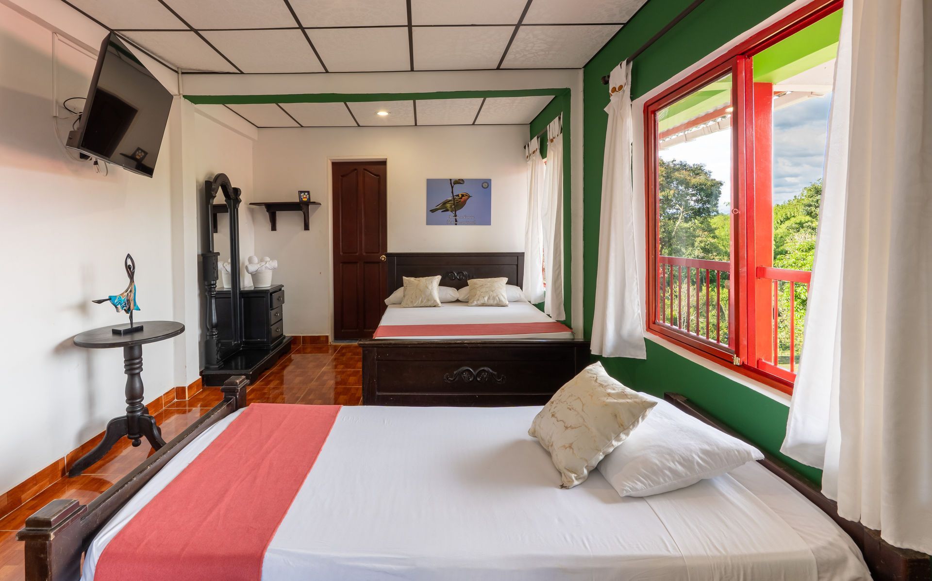 Habitación luminosa con dos camas, ventanas con marcos rojos y vistas al balcón a través de cortinas transparentes.