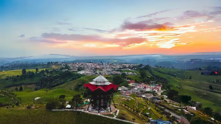 Amanecer sobre un pueblo verde en la ladera de una colina con una torre de techo rojo y campos ondulados.