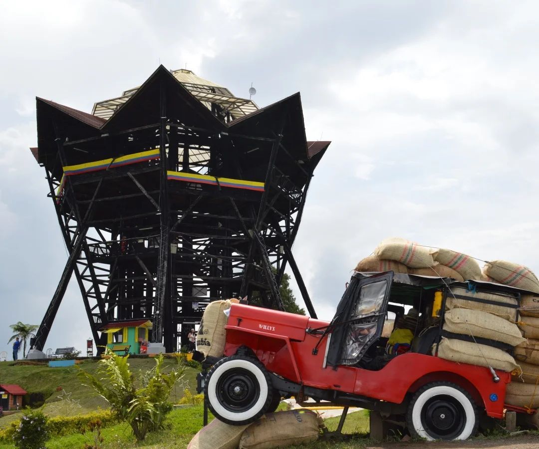Jeep rojo con sacos frente a una gran torre de observación negra bajo un cielo nublado.