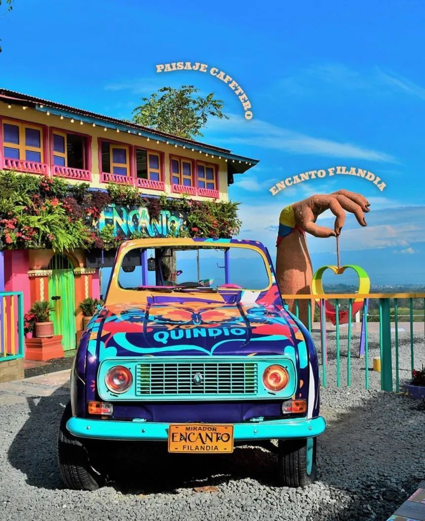 Camión antiguo y colorido frente a un edificio brillante, con una gran estatua de dinosaurio y un paisaje costero de fondo.