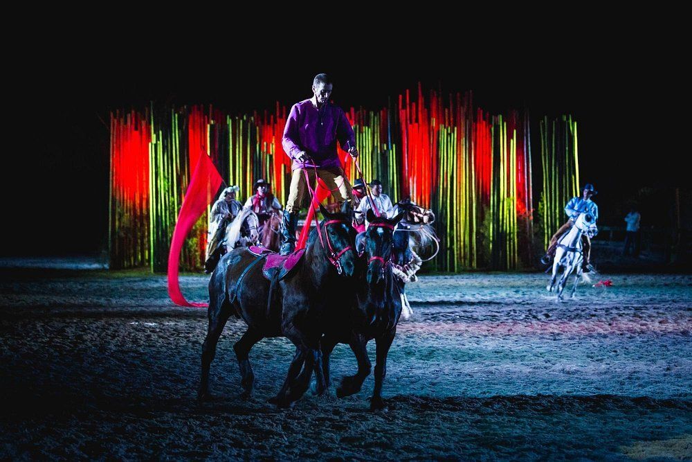 Artistas a caballo en una arena oscura con estelas de luces rojas y verdes brillantes a sus espaldas.