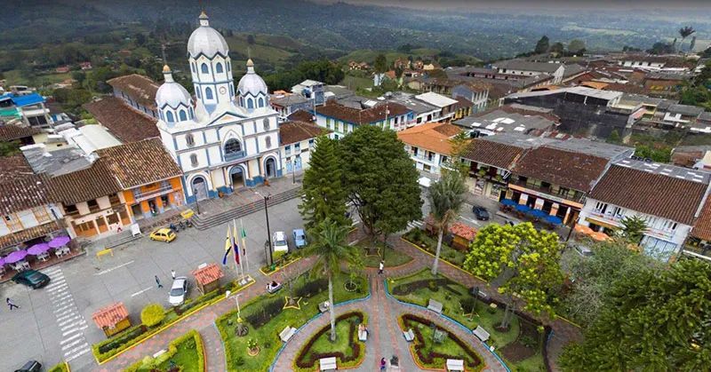 Vista aérea de una iglesia blanca junto a una plaza y jardines, con edificios de tejados de tejas y montañas al fondo.
