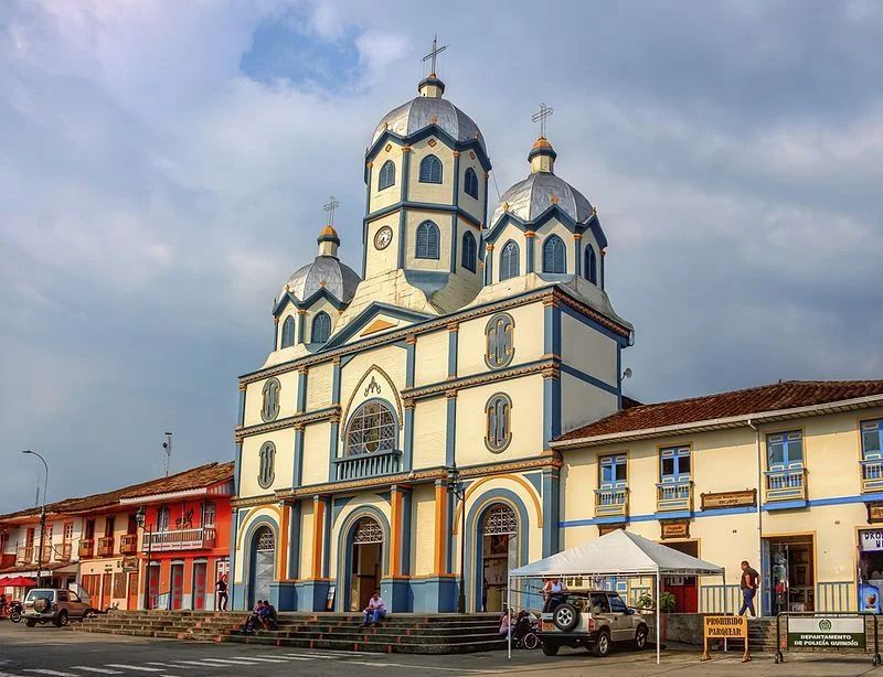 Iglesia azul y blanca con cúpulas en una plaza del pueblo, junto a coloridos edificios bajo un cielo nublado.