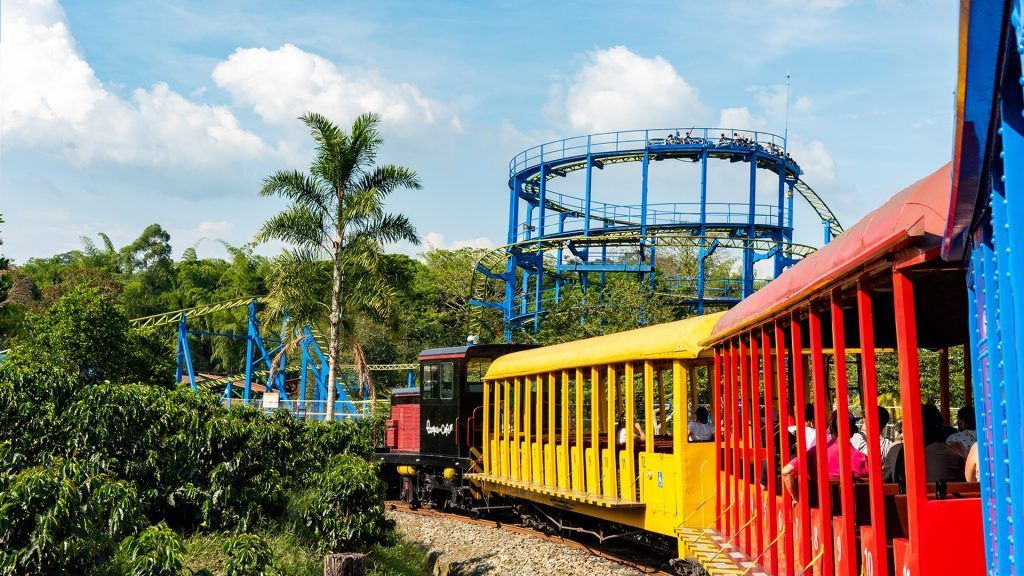 Vagones de tren de colores serpentean a través de un frondoso parque junto a una montaña rusa azul bajo un cielo brillante.