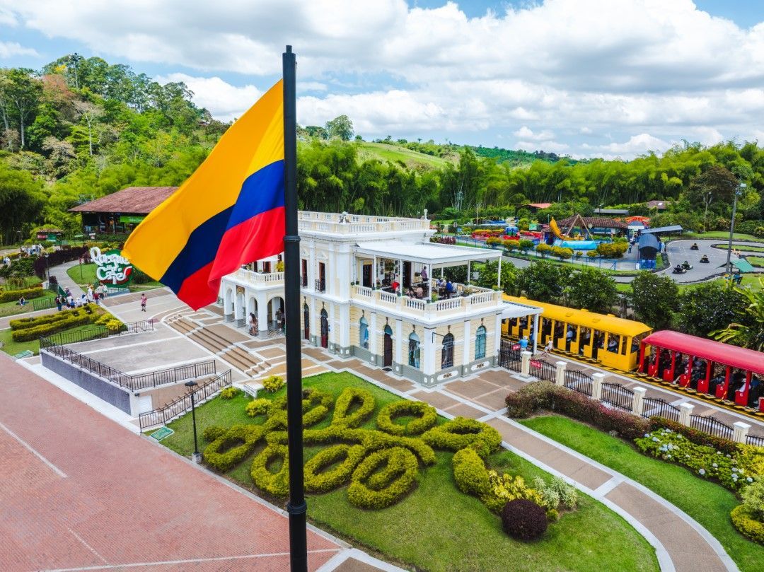 Bandera colombiana sobre un edificio colonial blanco con jardines paisajísticos y coloridos vagones de tren en las cercanías.