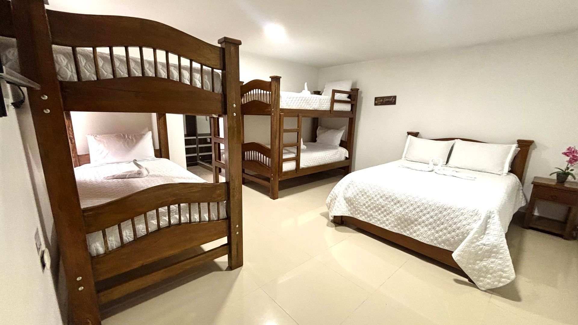 Habitación pequeña con literas de madera, una cama doble y ropa de cama blanca sobre suelo de baldosas.