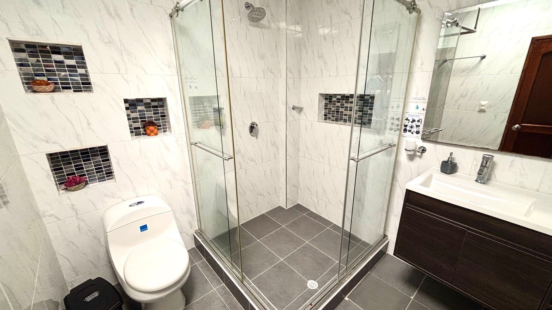 Baño moderno con ducha de cristal, inodoro blanco y tocador oscuro con espejo.
