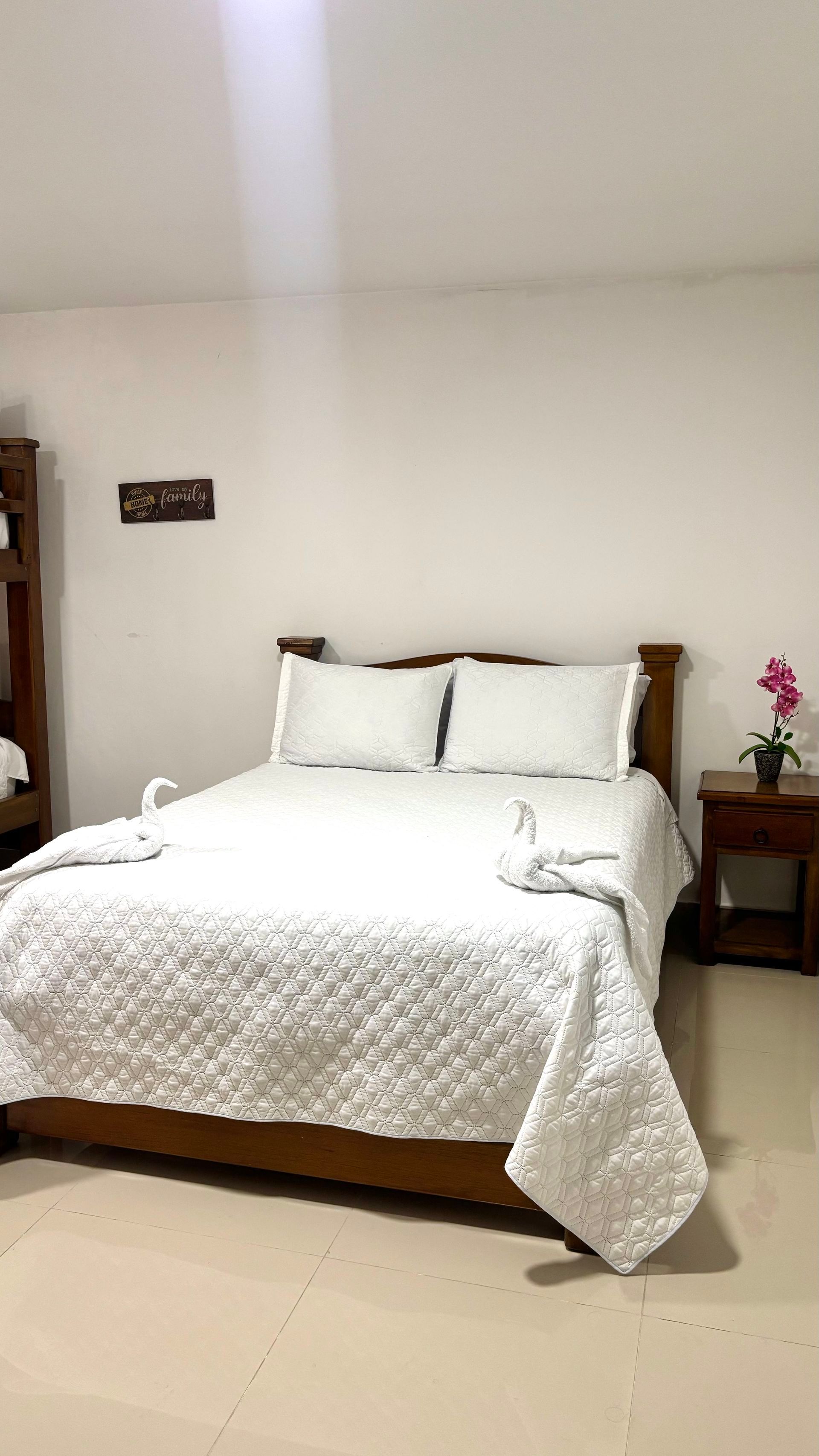Una cama blanca bien hecha con almohadas en un dormitorio sencillo, junto a una mesita con una flor rosa.