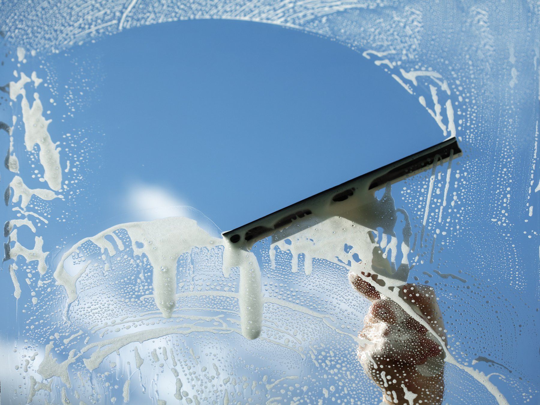 Person cleaning a window with a squeegee and soapy water against a blue sky.