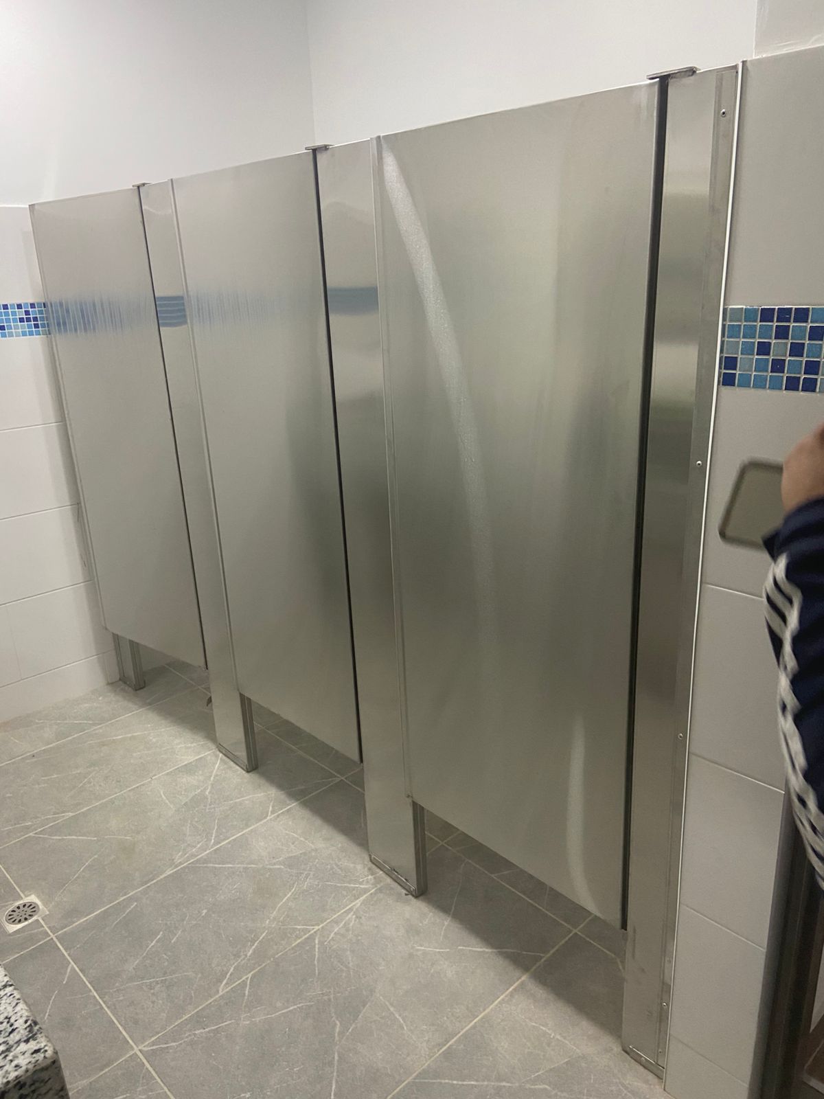 Tres cabinas de baño de acero inoxidable en un baño público con piso de baldosas grises y paredes blancas.