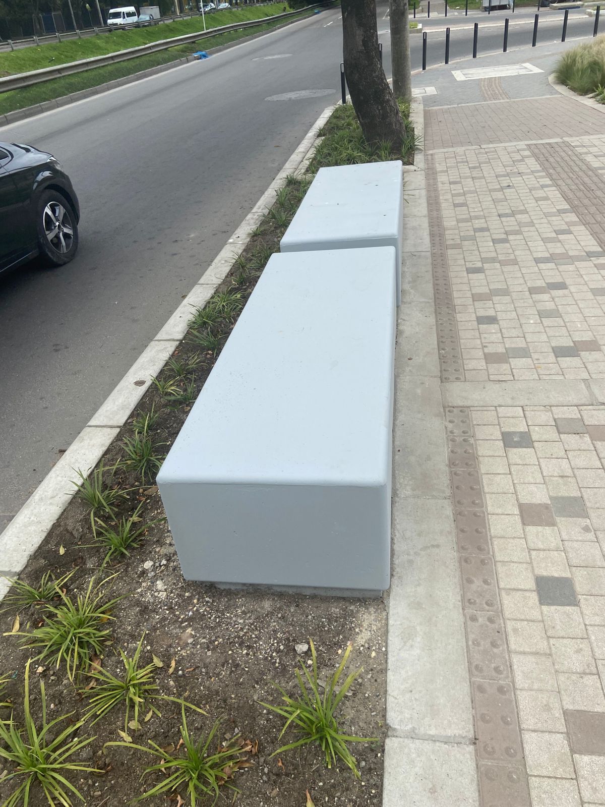 Dos bancos grises en una acera junto a una carretera, con un coche en movimiento.