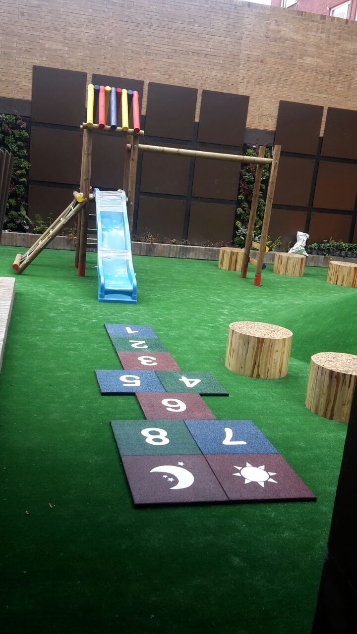 Parque infantil con tobogán azul, rayuela y columpios sobre césped verde.