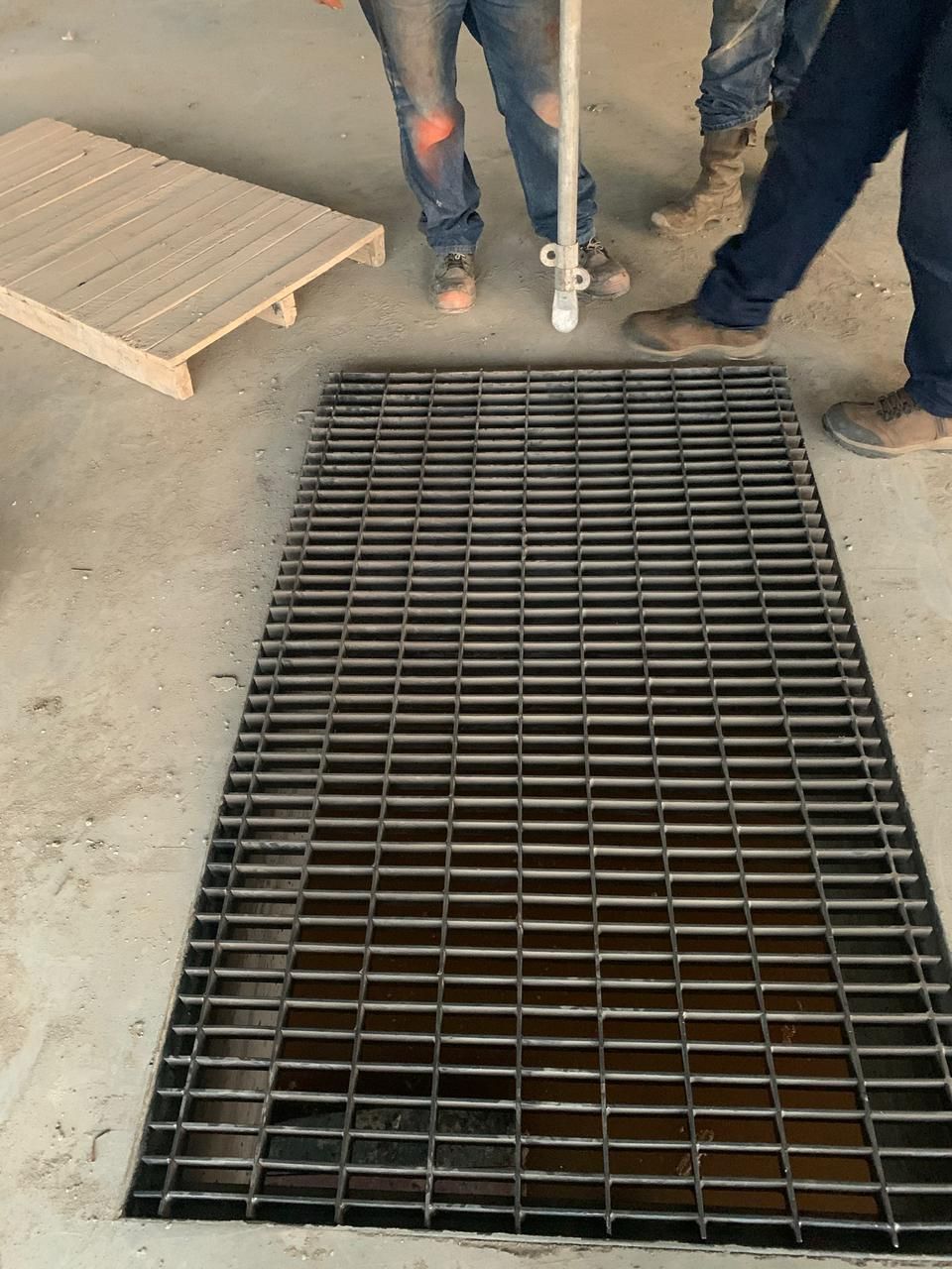 Trabajadores cerca de una rejilla de metal colocada en un piso de concreto, posiblemente cubriendo un punto de acceso.