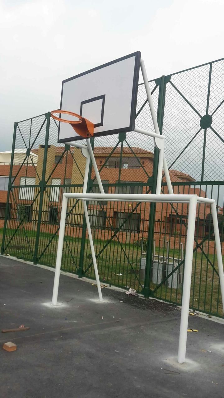 Aro de baloncesto blanco sobre una cancha negra. Valla verde y edificios al fondo. Cielo nublado.