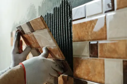A worker in work gloves installs brown and cream decorative tiles onto a wall covered with combed thin-set adhesive.