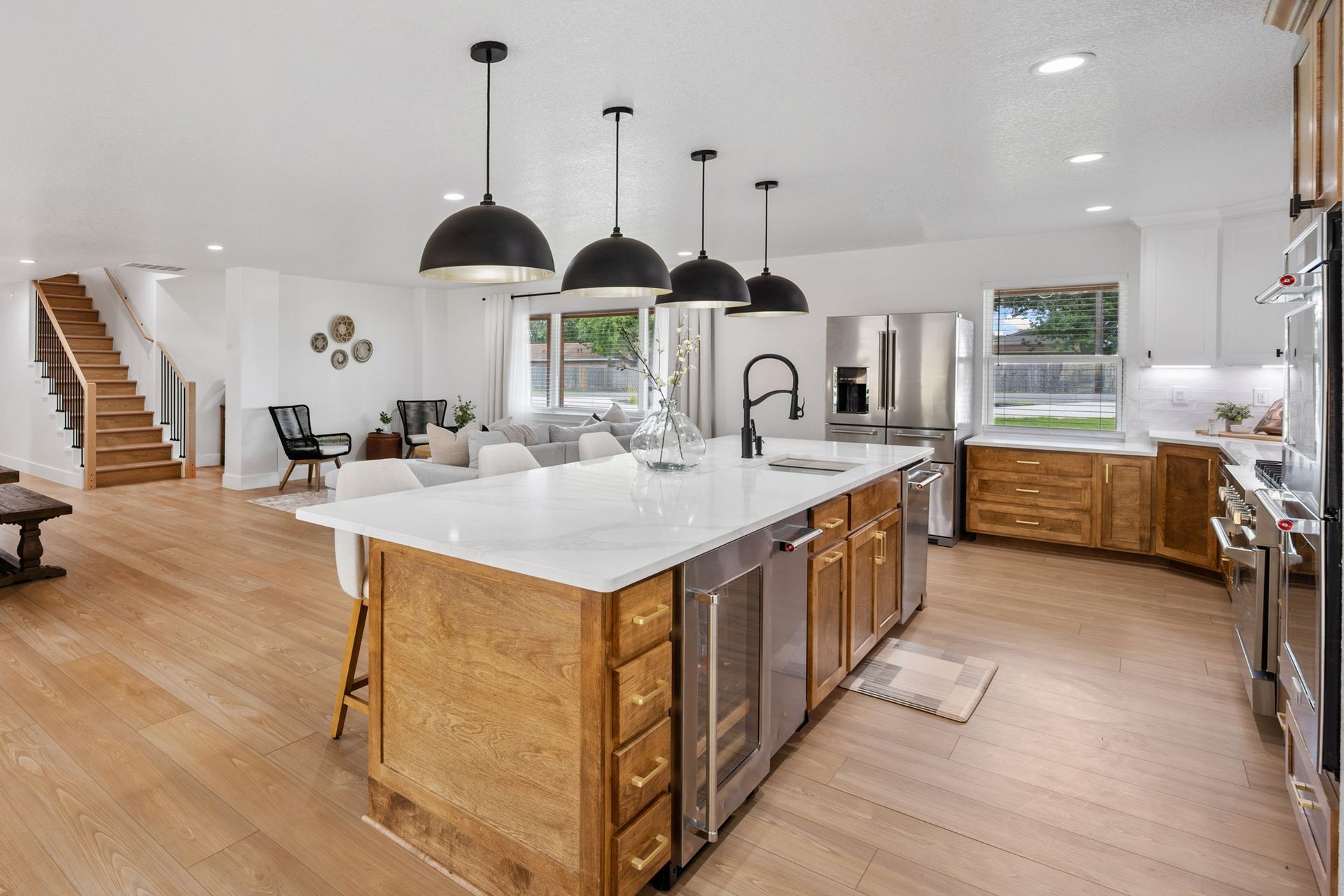 A bright, modern open-concept kitchen with wooden cabinets, a large white island, pendant lights, and a wooden staircase.