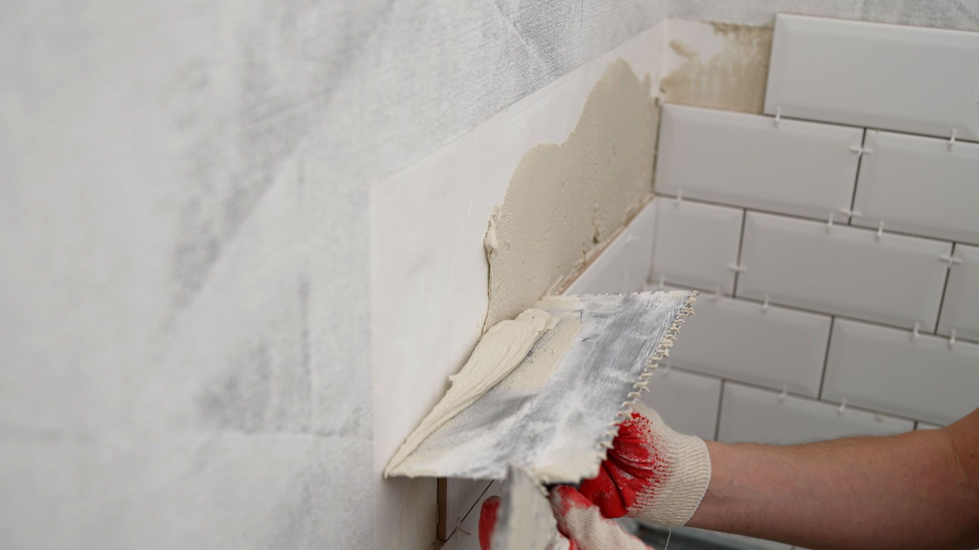 A gloved hand uses a trowel to apply thin-set mortar to a wall corner before installing white subway tiles.