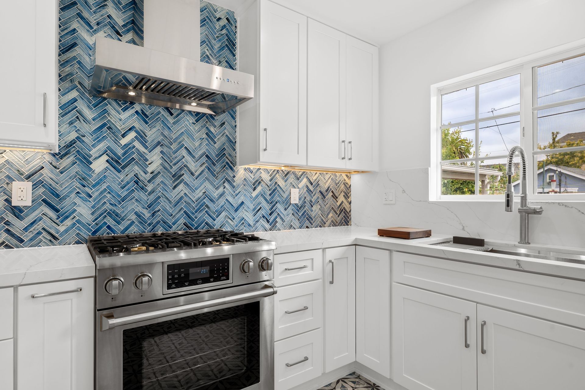 Modern white kitchen with stainless steel oven, blue herringbone backsplash, and a window above the sink.