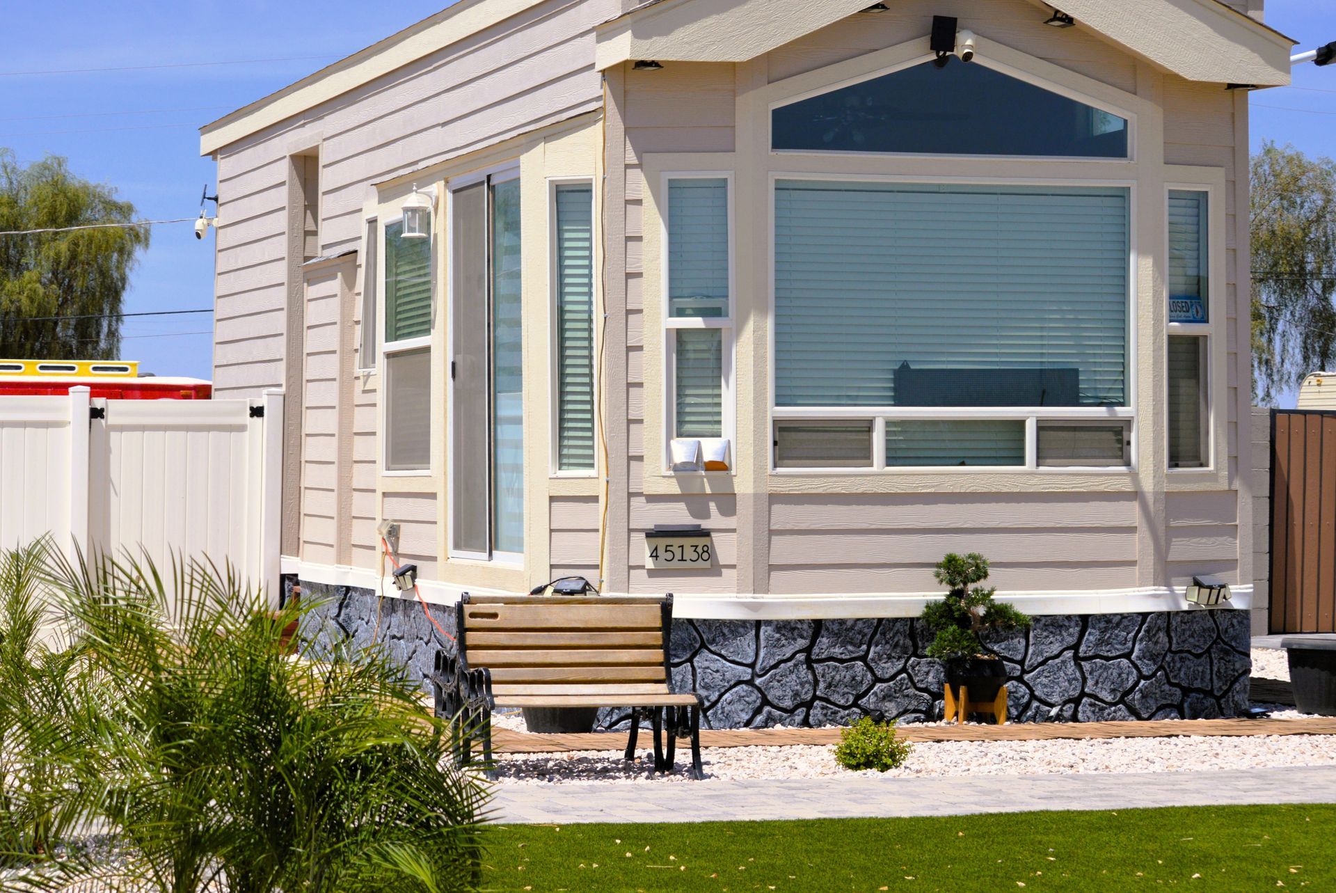A beige mobile home with a large front window, a wooden bench, and faux-stone skirting set on a paved lot with landscaping.