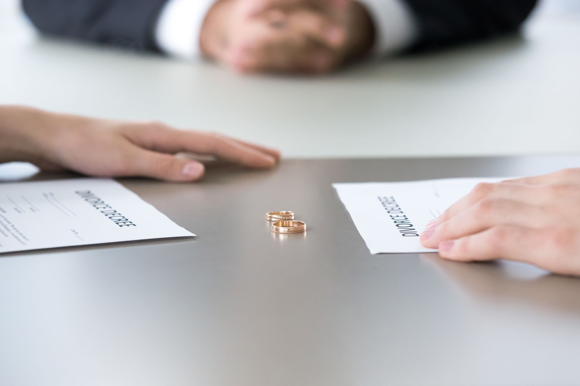 Two hands reach for divorce papers, wedding rings between them, a suited person in the blurred background.