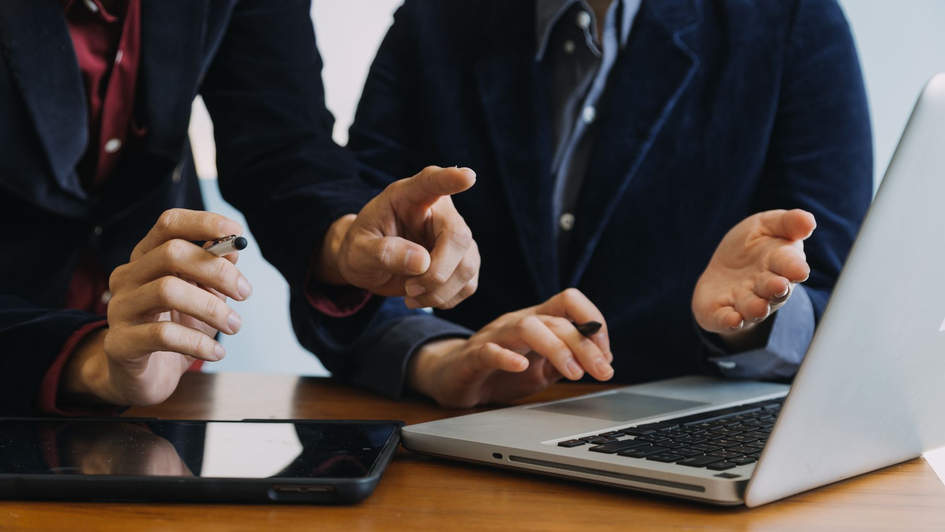 Two people in business suits pointing at a laptop and tablet on a wooden desk.